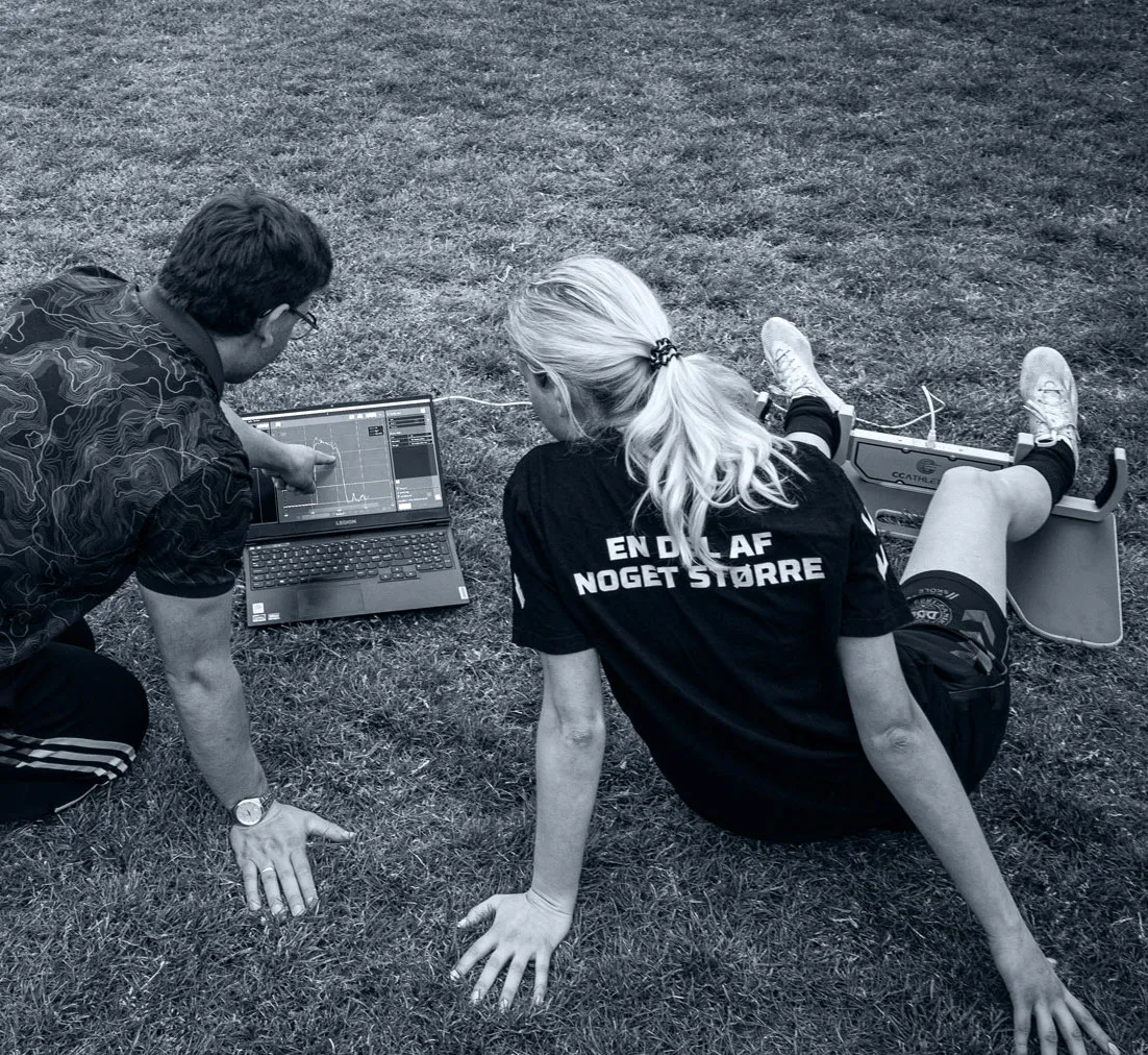 A coach reviews GroinMate results on a laptop while an athlete performs an adductor strength test on a grass field